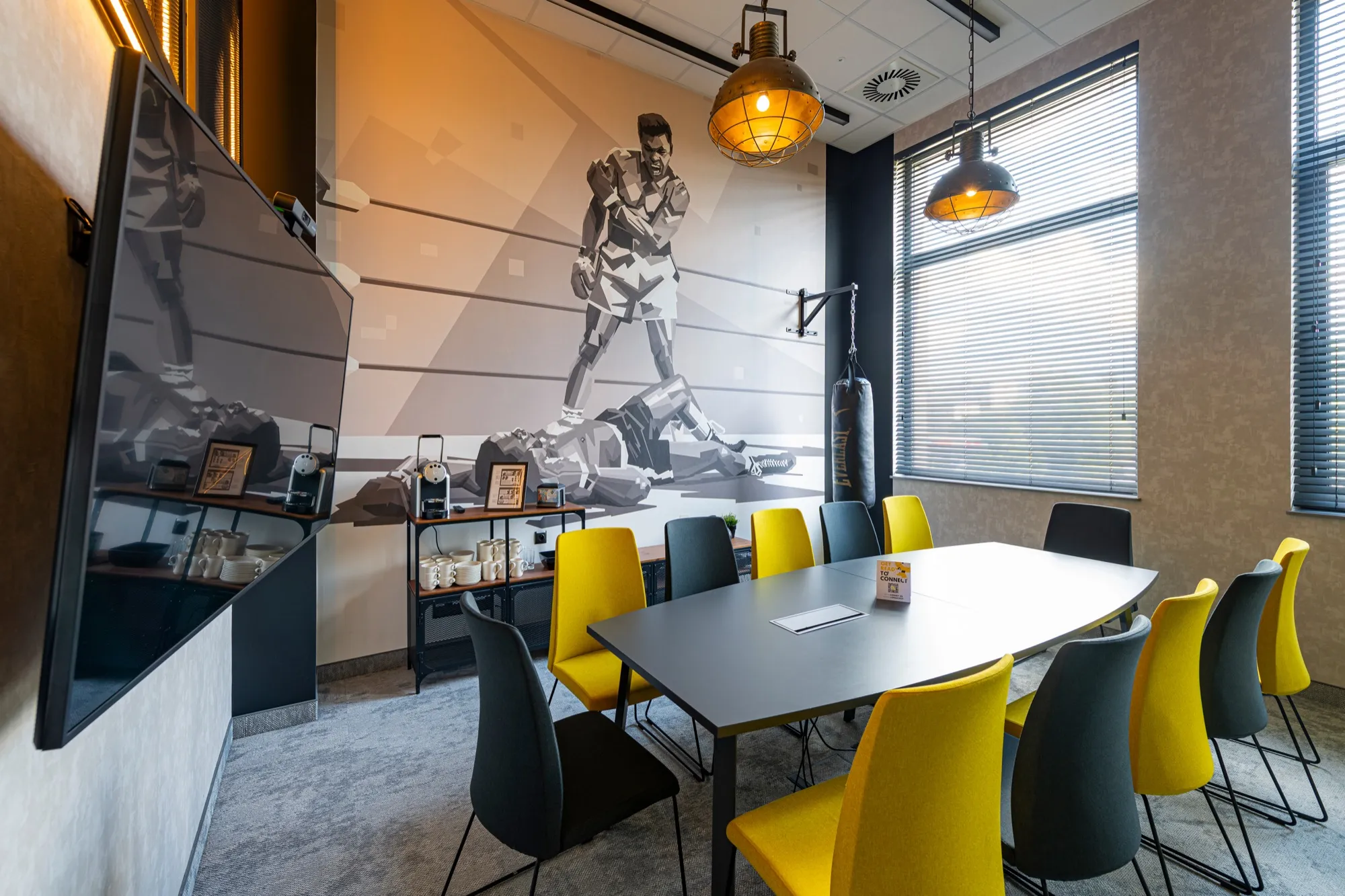 Sala kameralna w o3meet — stół konferencyjny, ekran, duże okna z żaluzjami i mural o tematyce sportowej